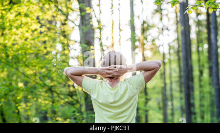 Junger Mann die Natur genießen, im Wald mit die Arme hinter dem Kopf nach der glühenden Sonne morgens durch die Bäume. Stockfoto