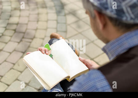 Zurück über die Schulter zu sehen, was der Mann mit dem Hut gekreuzten Beinen sitzen und ein Buch lesen. Stockfoto