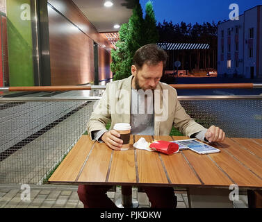 Ein bärtiger Mann mittleren Alters der kaukasischen Aussehens sitzt an einem Tisch in einem Sommer Cafe am Abend Aufpassen der Nachrichten auf dem Bildschirm eines Tablet, trinken Kaffee und essen Pommes frites. Stockfoto