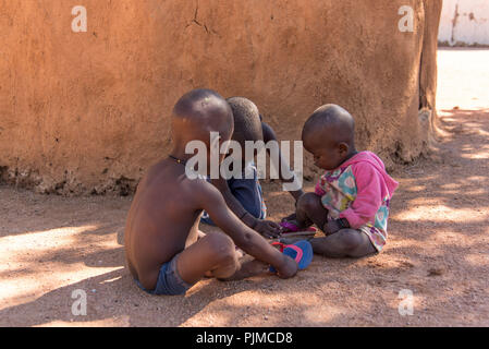 Drei Himba Kinder spielen in den Schatten einer Lehmhütte Stockfoto