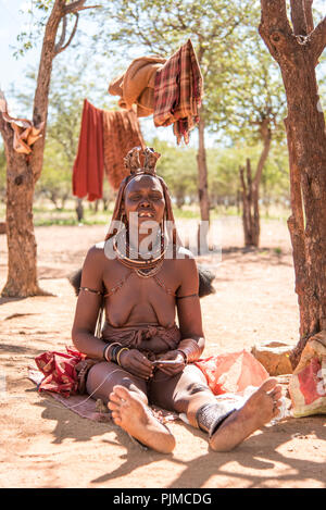 Frau Himba sitzt auf dem Boden und macht Schmuck. Man kann deutlich sehen, dass die mittleren Zähne - aus Tradition - entfernt wurden Stockfoto