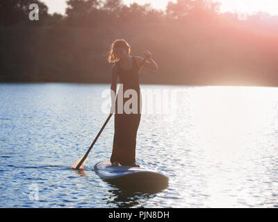 Silhouette einer schönen Frau in einem schwarzen Kleid am Stand up Paddle Board, Sonnenuntergang. SUP Stockfoto