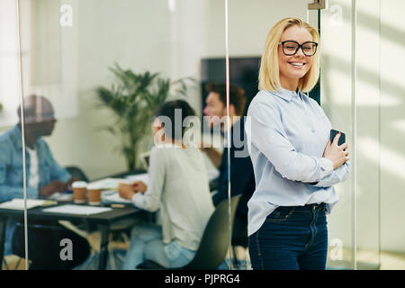 Lächelnden jungen Geschäftsfrau Brille trägt und Ihr Handy Holding beim Stehen an der Tür und ein Büro Boardroom mit Kollegen arbeiten in der Stockfoto