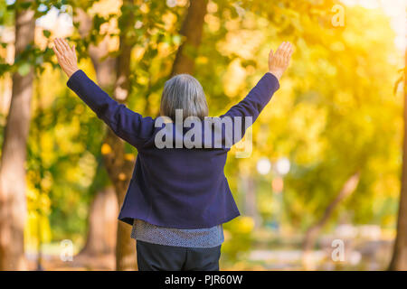 Casual ältere Frau mit ausgestreckten Armen stehen im Herbst Park. Rückansicht der älteren Frau, die Bäume im Herbst mit den Händen nach oben. Stockfoto