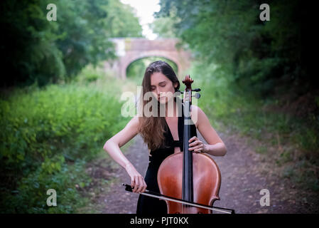 Eine junge blonde Frau, spielt Cello Draußen, hinter ihr, über einen Pfad eine Brücke aus gesehen werden kann. Stockfoto