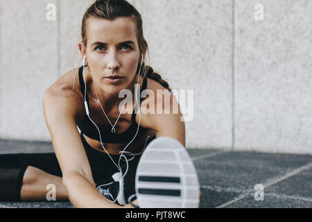 Fitness-Frau beugt sich nach vorne und dehnt ihr Bein auf dem Boden. Frau, die Dehnungsübungen macht und Musik hört, mit Ohrhörern sitzend Stockfoto