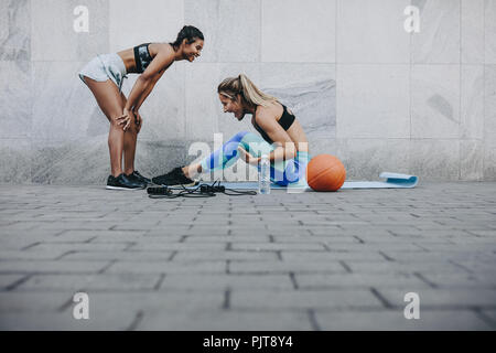 Seitenansicht der Frau tun, Sit Ups, während ihr Freund entspannt mit den Händen auf den Knien. Zwei fitness Frauen in fröhliche Stimmung beim Training im Freien. Stockfoto