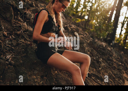 Junge Frau sitzend durch Mountain Trail prüfen Ihr Smartphone. Frau ausruhen nach Trail laufen und prüfen Ihr smart Watch. Stockfoto