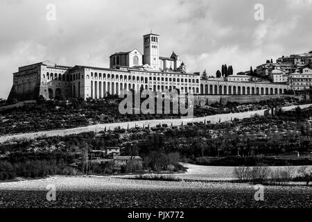 Auf Assisi Stadt und St. Franziskus Kirche (Umbrien) im Winter, mit einem Feld durch Schnee und Himmel mit weißen Wolken bedeckt Stockfoto