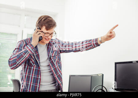 Zornigen jungen Mann sprechen auf dem Mobiltelefon und empfängt fehlerhafte Nachrichten während Stading an seinem Arbeitsplatz im Büro. Stress Gespräch. Stressige Situationen Stockfoto