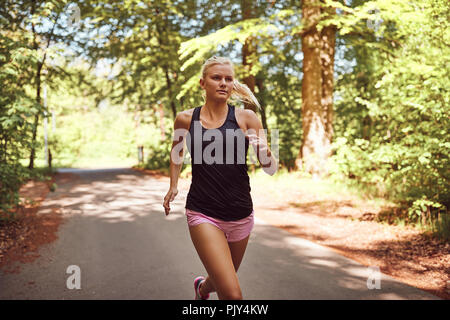 Passen junge blonde Frau in Shorts und einem Tanktop joggen allein auf einem Pfad durch den Wald an einem sonnigen Tag Stockfoto