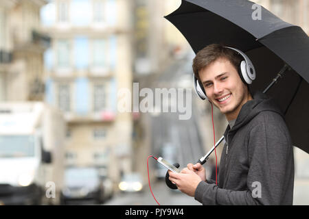 Happy Teen Musik hören mit Kopfhörern an Suchen Sie auf der Straße unter dem Regen Stockfoto