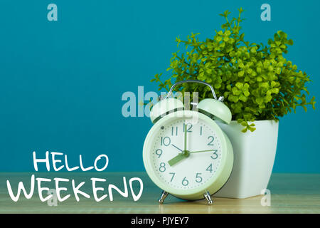 Hallo Wochenende Text auf grünem Hintergrund und kleinen Baum in weiße Vase mit Vintage Wecker auf Holztisch. Stockfoto