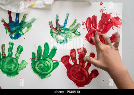 Blick von oben auf die eine Hand von einem Kind, das Rot und Grün bunte Palm druckt auf ein weißes Stück Papier. Detailansicht. Stockfoto