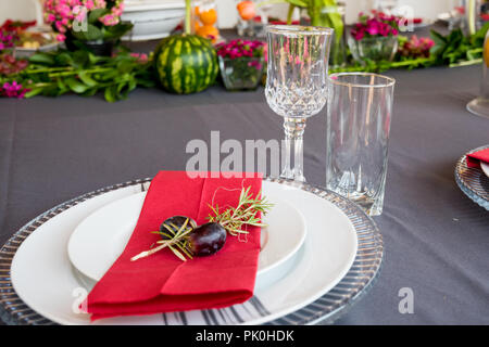 Ein Mittagstisch mit unter Teller Set, schöne graue und weiße Platten, rot Serviette, kleine dekorative tree branch, zwei Trauben mit Blumen und Obst Dekor. Stockfoto