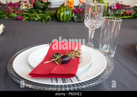 Ein Mittagstisch mit unter Teller Set, schöne graue und weiße Platten, rot Serviette, kleine dekorative tree branch, zwei Trauben mit Blumen und Obst Dekor. Stockfoto