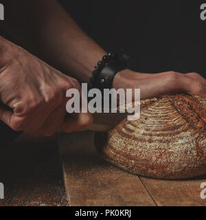 Close-up auf einem alten, braunen Tisch, ein Mann schneidet eine Runde dunkles Brot auf einem Holzbrett vor einem dunklen Hintergrund Stockfoto