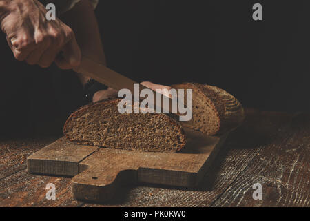 Männer Hände hausgemachte ein frisch gebackenes Brot auf einem hölzernen Schneidebrett auf dem Hintergrund eines alten, braunen Tisch Stockfoto