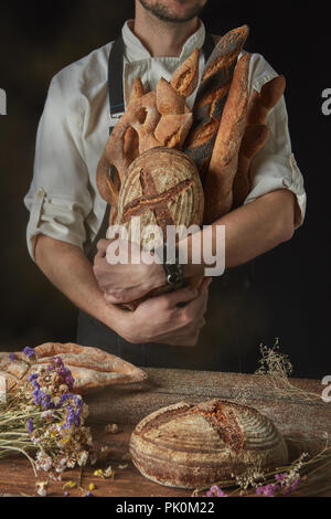 Auf einem dunklen Hintergrund ein Bäcker in einer schwarzen Schürze hält ein frisch gebackenes Brot und hölzernen Tisch mit Brot und getrocknete Blumen Stockfoto