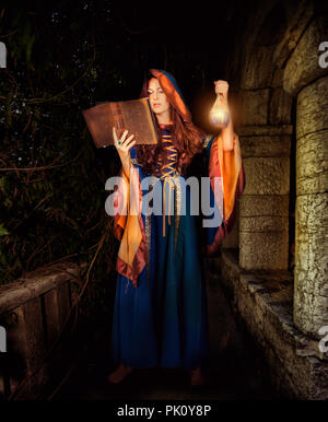 Schöne junge Halloween Hexe Vintage gothic Kleid mit Kapuze magische Buch von Zaubersprüchen in alten Lederbezug und trank in Glas halten Stockfoto