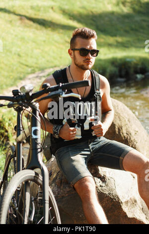 Stilvolle junger Mann mit einem Fahrrad auf einem Stein saß. Schwarzes T-shirt mit Print 23. Stockfoto