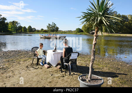 09.09.2018, Sachsen-Anhalt Wörlitz: Auf der Sandbank an der See im Wörlitzer Gartenreich, sonst von über einem Meter Wasser bedeckt, eine Frau und ein Kind sitzen an einem kleinen Tisch. Die GONDOLIERI, die nicht mehr in der Lage sind, die Kanäle mit den Besuchern zu navigieren, kann aber Teil des Sees verwenden, möchten Sie zu verschönern, die Ausgetrockneten Randgebieten mit Ihrer Installation. Die Dürre, die für die Monate gedauert hat, abgelaufen ist, fast die gesamte umfangreiche Kanalsystem im Wörlitzer Gartenreich, das zum Weltkulturerbe der UNESCO gehört. Der landschaftspark zwischen Dessau-Roßlau und Wittenberg war ou festgelegt Stockfoto