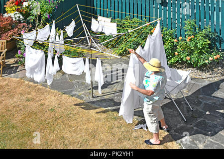 Hot 2018 Sommer auf geröstete trocken zurück Garten Rasen & reife ältere Frau mit Sonnenhut hängenden Waschen bei Rotary Wäscheleine England Großbritannien Stockfoto
