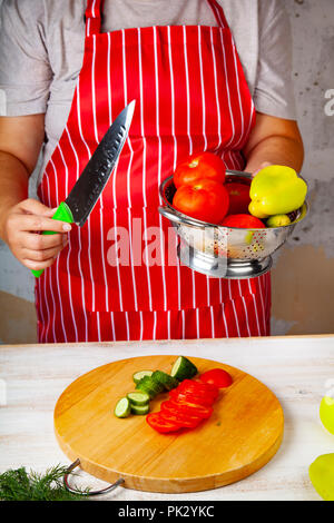 Frau in einem roten Schürze mit einem Sieb mit Gemüse für Salat und Messer. Das Kochen. Stockfoto