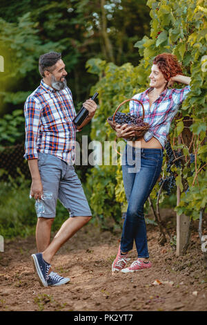 Schöne lächelnde Paare sind Spaß und einer Weinprobe auf einem Weingut. Ein Mann hält eine Flasche Wein und eine Frau hält Korb mit Trauben. Stockfoto