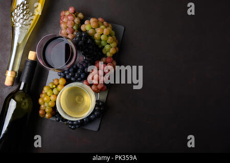 Zwei Weingläser mit Rot- und Weißwein, Flaschen und Trauben auf dunklem Hintergrund. Ansicht von oben. Flach. Platz kopieren Stockfoto