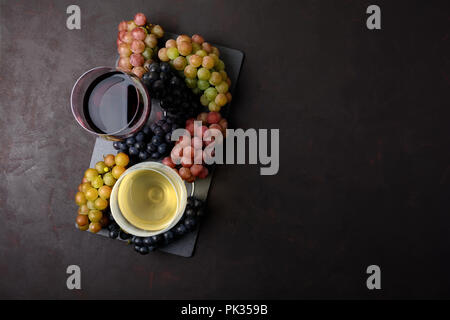 Zwei Weingläser mit Rot- und Weißwein, Flaschen und Trauben auf dunklem Hintergrund. Ansicht von oben. Flach. Platz kopieren Stockfoto