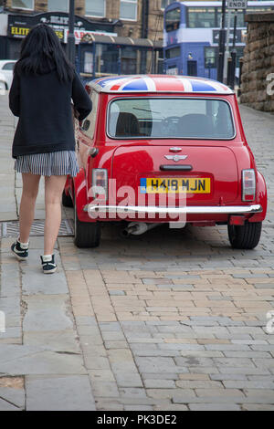 Rückansicht eines Roten Mini Cooper mit einem Union Jack Dach mit einem Mini skirted Mädchen zu Fuß mit dem Auto. Ein John Cooper Version des Autos auf einer Straße geparkt Stockfoto