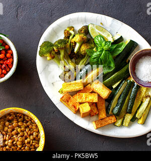 Gesunde vegetarische Nahrung. Gebackene Gemüse (Zucchini und Broccoli), gebratener Tofu, Käse, Braten, Kichererbsen und grüne Linsen auf dunklem Hintergrund, Ansicht von oben Stockfoto
