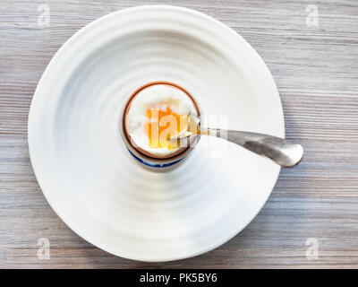Blick von oben auf die weich gekochten braunen Ei mit Löffel in der Tasse auf weiße Platte auf grau Holzbrett Stockfoto