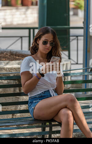 Attraktive junge Frau mit schönen Beinen und mit Sonnenbrille und Shorts benötigten ein Smartphone sitzt in der Sonne. Stockfoto