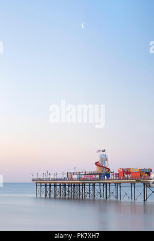 Mond über steigende in Torbay Paignton Pier wie die Farben des Sonnenaufgangs burst über den Himmel und das Meer ist ruhig. Stockfoto