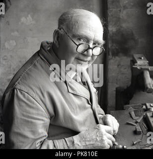 1950, historische, eine ältere männliche Handwerker in einem traditionellen workcoat oder Overalls an seiner Werkbank Reparatur Werkstatt Werkzeuge sitzen, England, UK. Stockfoto