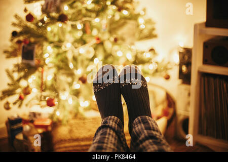 Festliche Socken auf alten hölzernen Stuhl auf dem Hintergrund der Goldenen schönen Christbaum mit Lichtern in festlichen Raum. Entspannen. gemütlichen Winterurlaub. warm Stockfoto