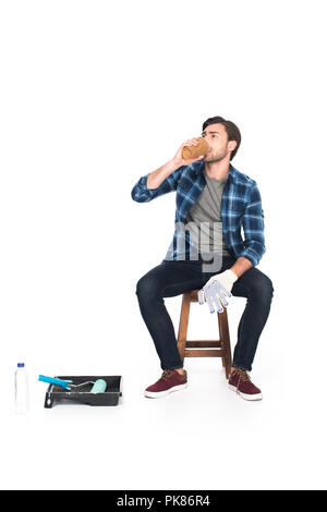 Man ruht auf einem Stuhl und trinkt Kaffee in der Nähe der Walze für Fach und Farbroller auf weißem Hintergrund Stockfoto