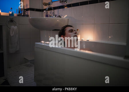 Junge Mann ist Entspannung in der Badewanne mit einer Maske auf und Kerzen um ihn herum leuchtet. Stockfoto