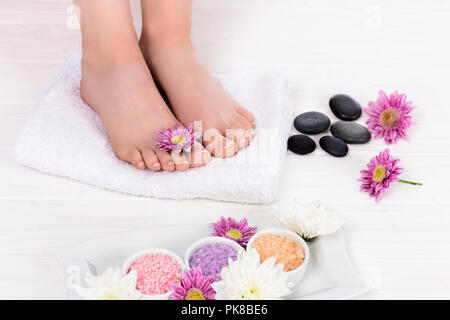Zugeschnittenes Bild der Barefoot Frau auf Spa-behandlungen mit Handtuch, Blumen, bunt Meersalz und spa Steine Stockfoto