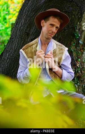 Ein Mann gekleidet wie ein Cowboy gegen einen Baumstamm Holding den Kragen seines Hemdes lehnt. Stockfoto