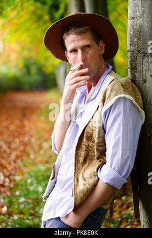 Ein Mann gekleidet wie ein Cowboy gegen einen Baum Rauchen einer Zigarette lehnt. Stockfoto