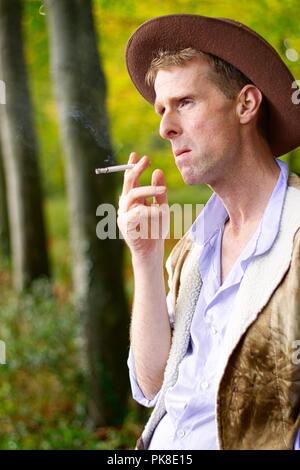 Ein Mann gekleidet wie ein Cowboy gegen einen Baum Rauchen einer Zigarette lehnt. Stockfoto