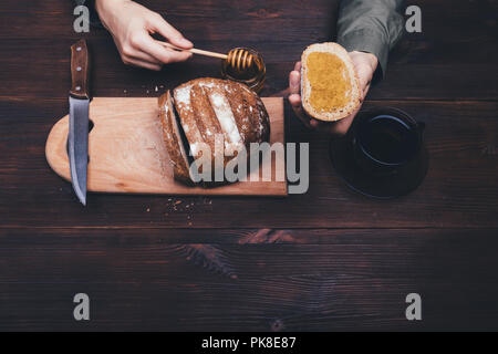 Ansicht von oben weibliche Hände hinzufügen Honig auf Roggen Sandwich auf dunklen Holztisch neben Tasse schwarzen Tee oder Kaffee. Zusammensetzung der Einfache rustikale Küche. Stockfoto