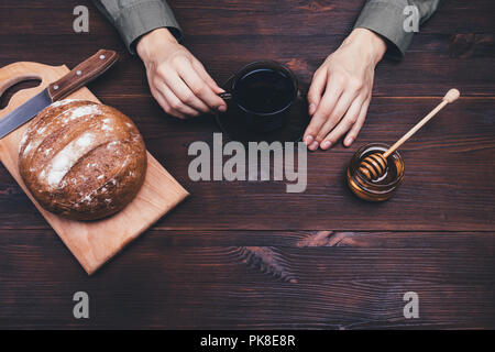 Weibliche Hände, die Tasse Tee oder Kaffee auf dunklen Holztisch, Ansicht von oben. Einfaches ländliches Frühstück mit Honig und Roggenbrot. Stockfoto