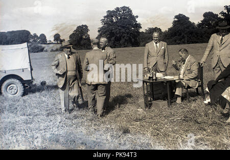 1930er Jahre, historischen, ländlichen Land Verfolgungsjagden, wel-gekleideten Herren auf geeignete Kleidung stand auf einem Hügel, durch eine Tabelle mit Trophäen mit dem Schießen Kapitän sitzen Rauchen einer Pfeife. Die Aufnahme ist eine traditionelle britische Landschaft fieldsport mit strengen Regeln auf Sportlichkeit und Etikette. Stockfoto