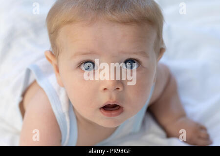 Niedlich neugierig Blauäugigen jungen Baby an der Kamera starrt, wie es auf den Bauch auf einer weißen Bettdecke auf einem Bett liegt Stockfoto