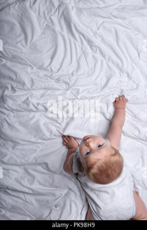 Neugierig cute baby baby boy Kriechen auf ein weißes Bett seinen Kopf zurück muss in die Kamera in einer Ansicht von oben mit Kopie Raum zu suchen Stockfoto