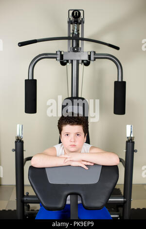 Cute fat wenig kaukasischen Junge sitzt auf Sport Übung Maschine in Fitness Gym im Innenbereich Stockfoto